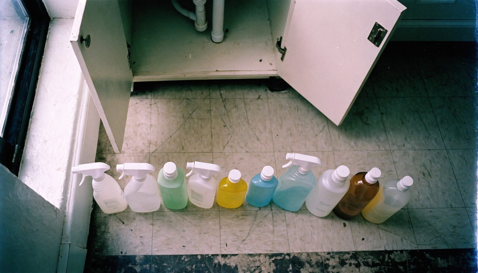 Eleven plastic spray bottles of household cleaner lined up in a row on a kitchen floor in front of an open under-sink cabinet.