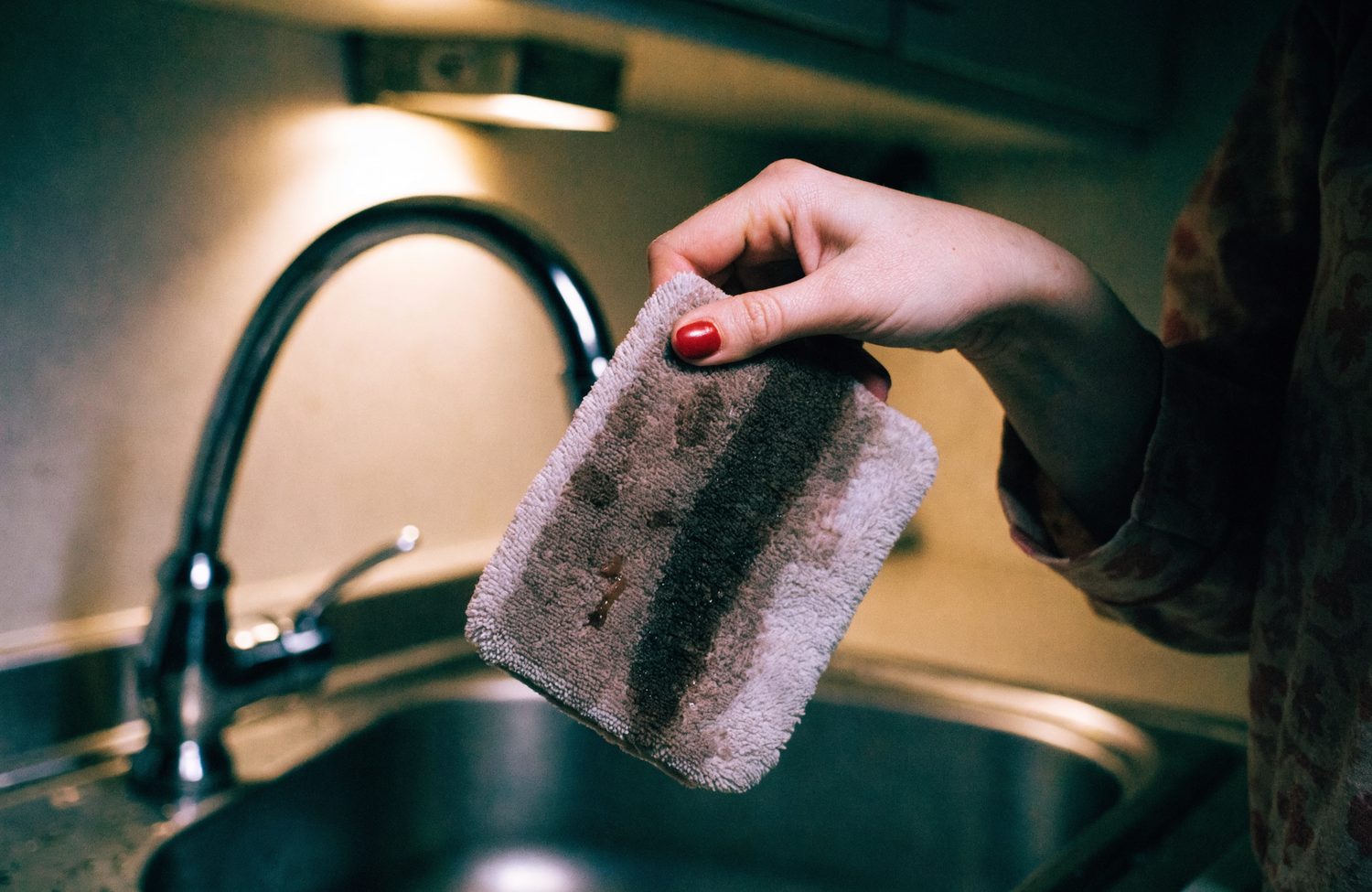 A hand holding up a used gray microfiber cleaning pad streaked with dark grime above a kitchen sink.