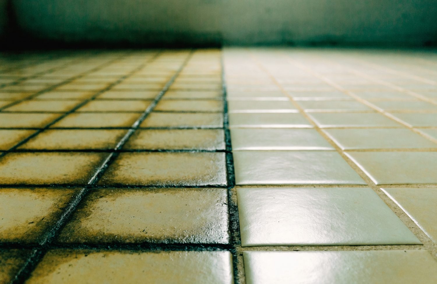 Close-up of an old tile bathroom floor with a straight dividing line: discolored grout on one side, clean pale grout on the other.