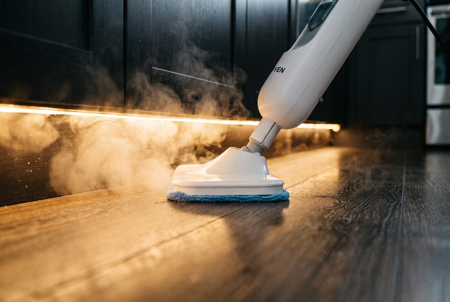The head of a steam mop pressed flat into the toe-kick gap beneath kitchen cabinets with steam curling out from underneath.